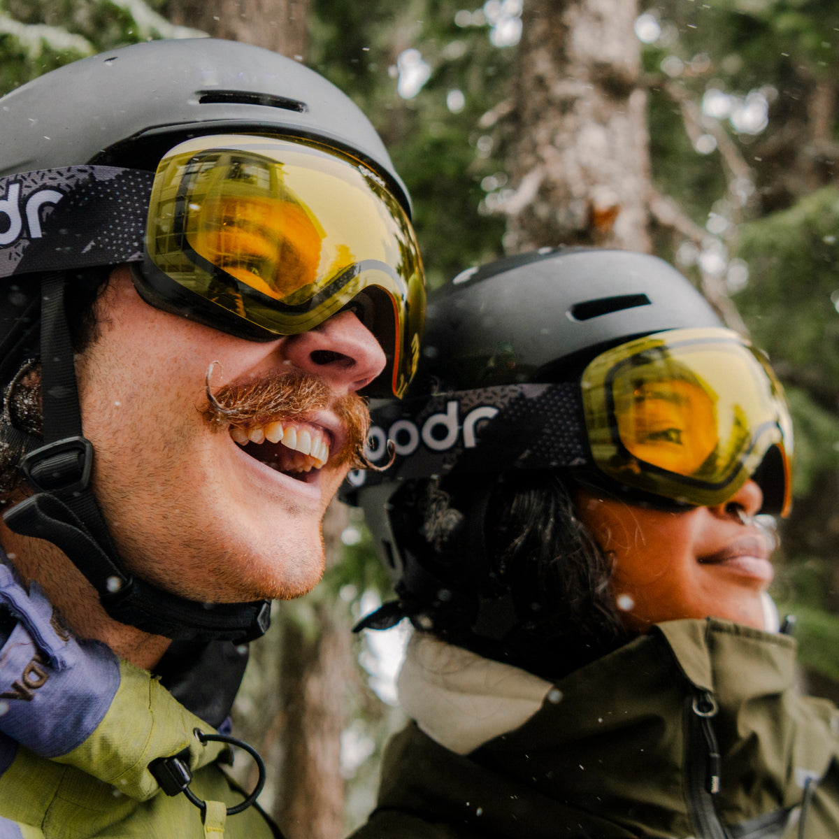 Two skiers wearing yellow snow goggles with patterned goodr straps and black helmets.