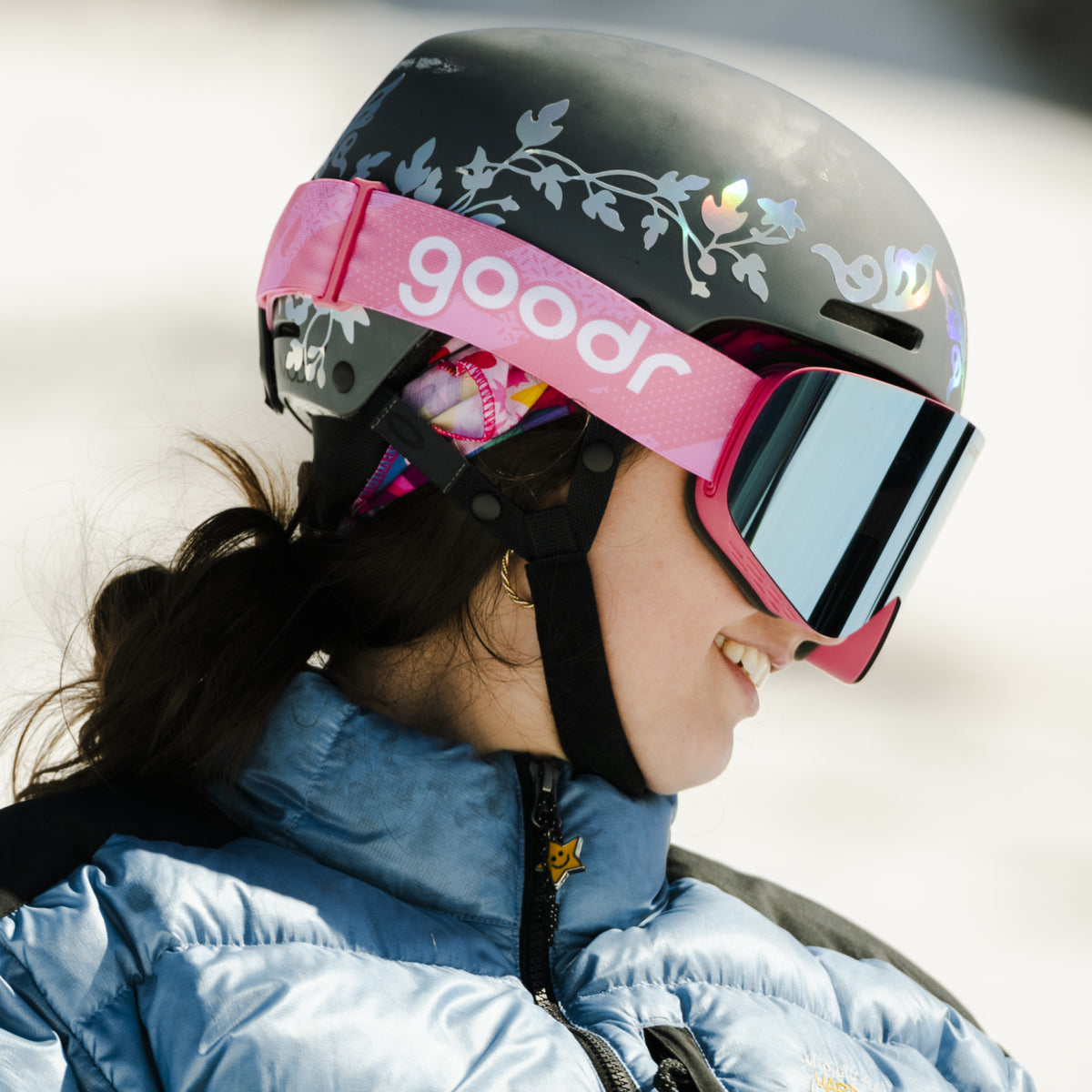 Skier wearing blue mirrored snow goggles with a pink goodr strap and black helmet.