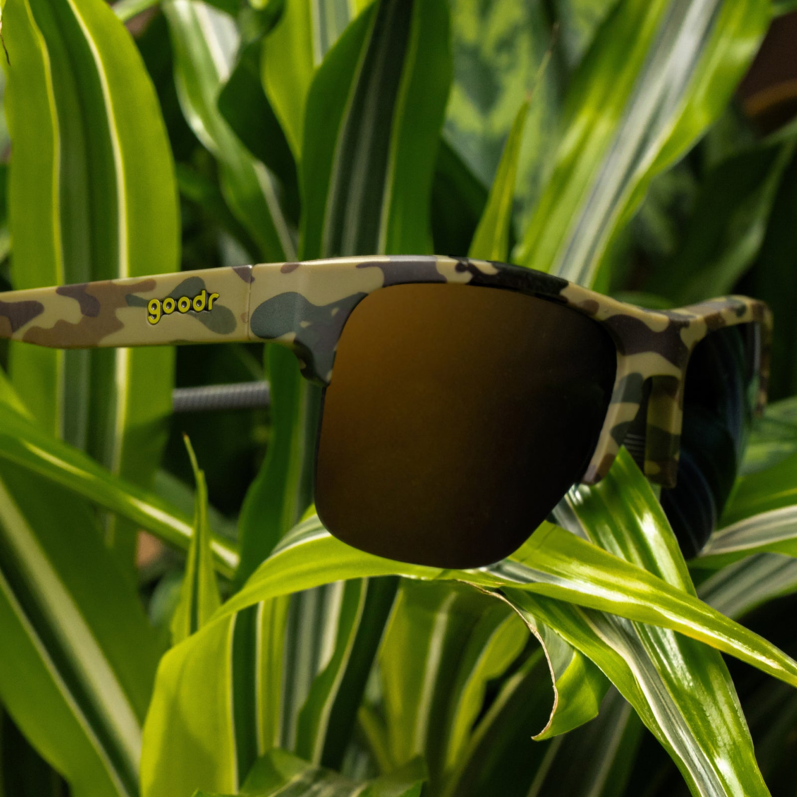 A pair of sunglasses with a green camouflage pattern frame and dark mirrored lenses, resting on green variegated leaves. The brand name 'goodr' is printed in yellow on the left temple arm.