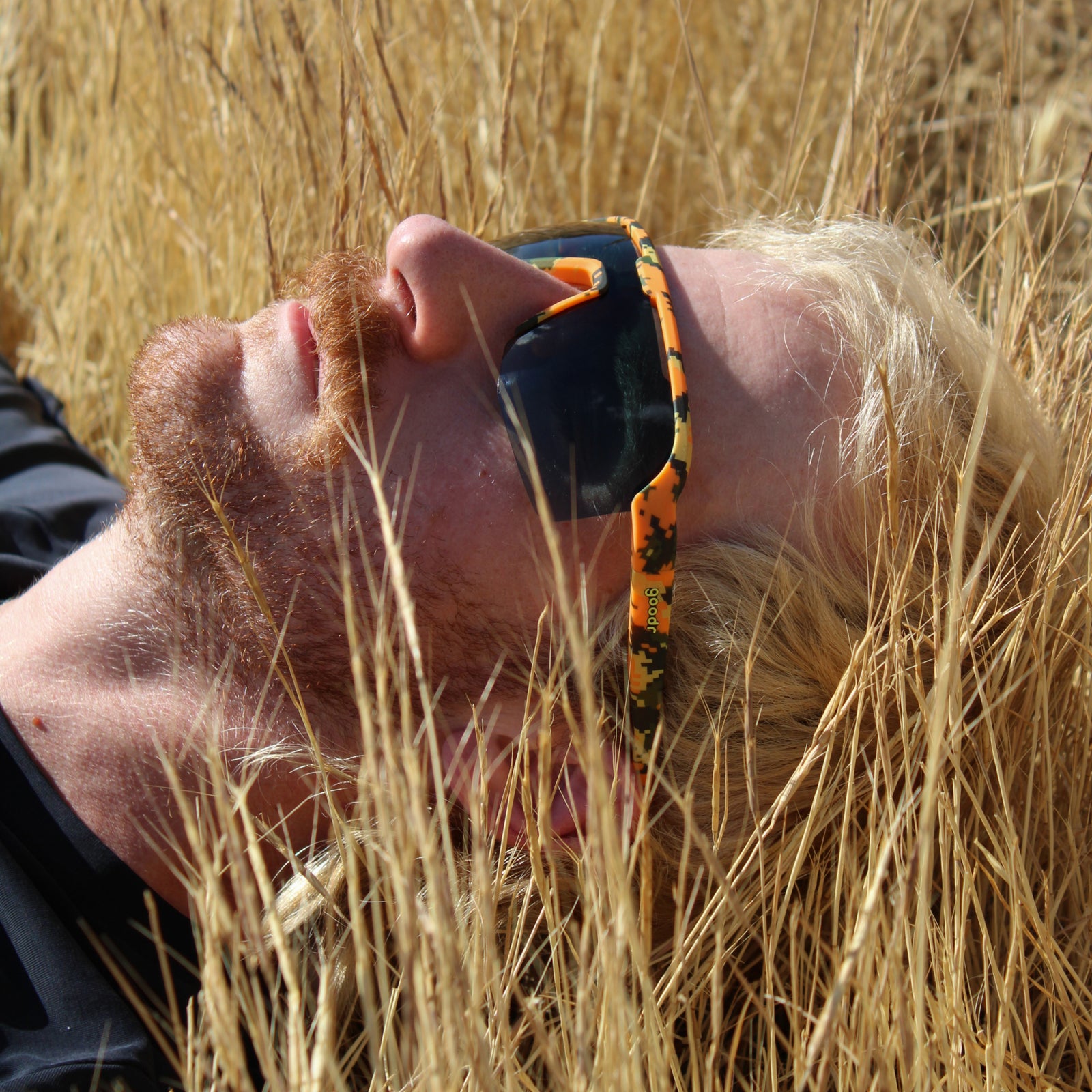 A close-up lifestyle shot of a man lying in a field of dry golden grass, wearing sunglasses with a vibrant orange and black patterned temple arm.