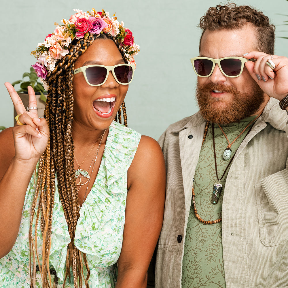 Woman wearing a floral crown and light green sunglasses, making a peace sign and laughing, next to a man with a beard wearing similar sunglasses, both dressed in earthy tones.
