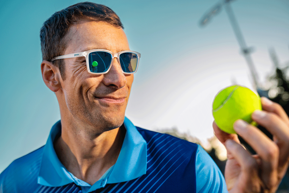 Man wearing white-framed sunglasses with reflective lenses, holding a tennis ball and smiling, dressed in a blue sports shirt under a clear sky.