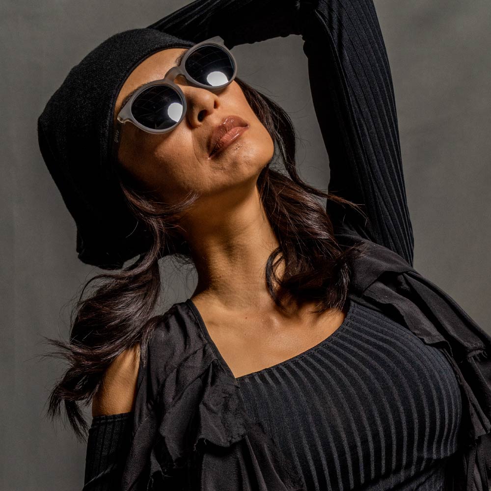 A woman wearing a black long-sleeved top, a black beret, and gray sunglasses with black lenses, posing with an arm over her head.