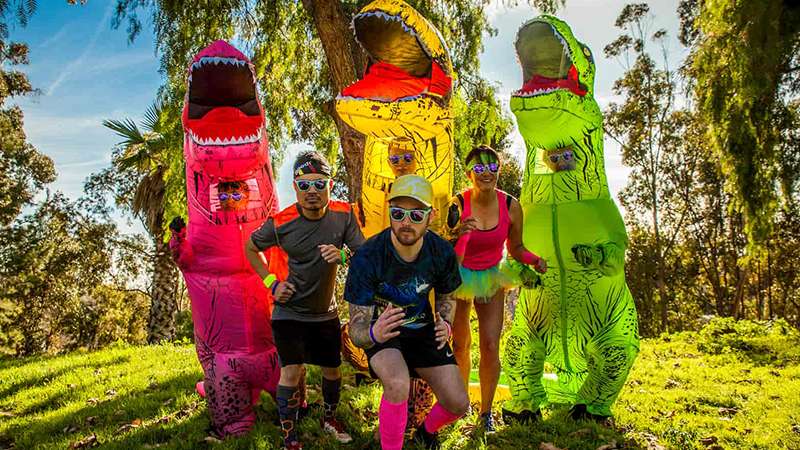 People wearing sunglasses in colorful inflatable dinosaur costumes standing in a sunny park.