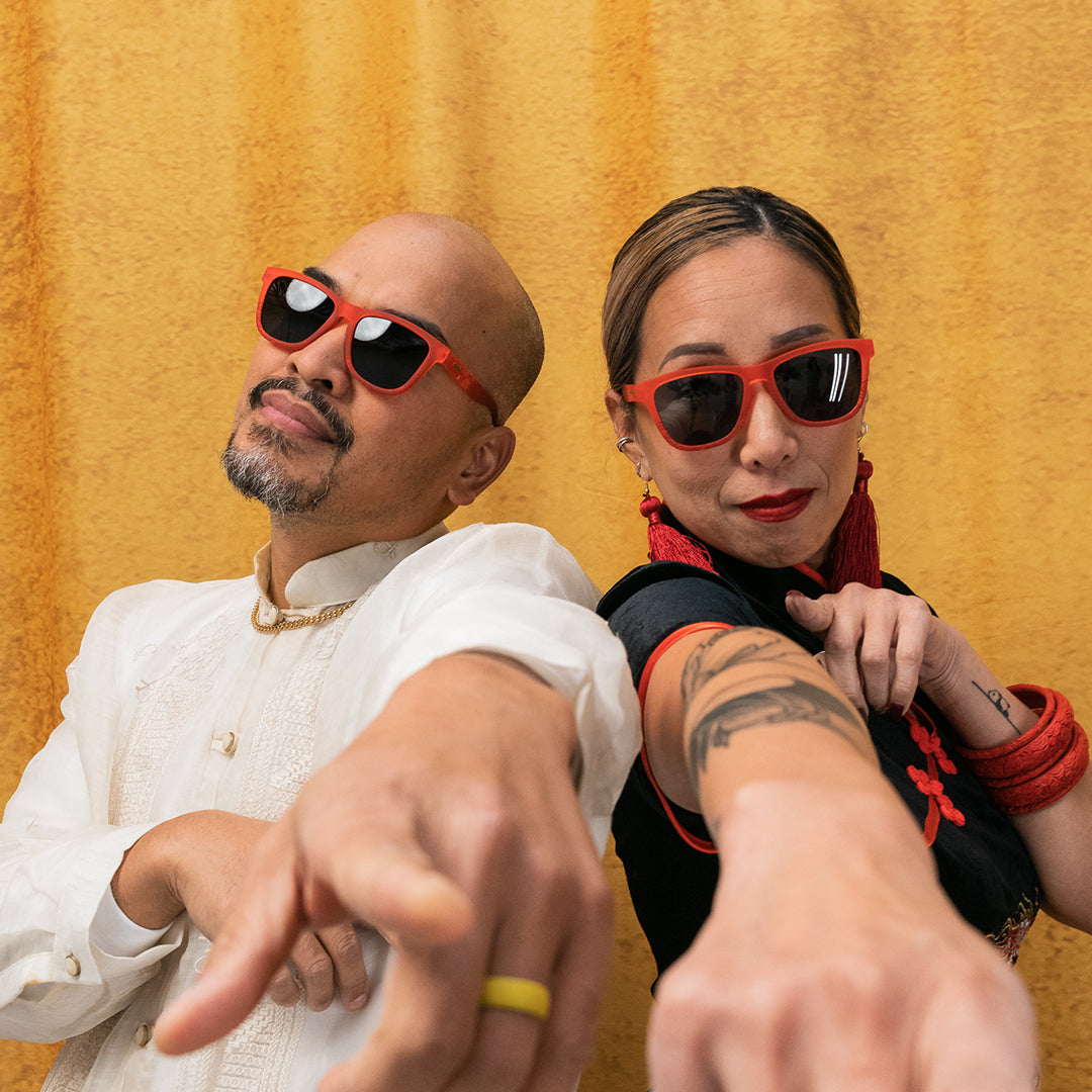 A man and a woman, both wearing red sunglasses, pose confidently, pointing toward the camera. The man has a shaved head, goatee, and wears a traditional white shirt, while the woman has dark red lipstick, large red earrings, and tattoos on her arm. A golden backdrop frames the shot.