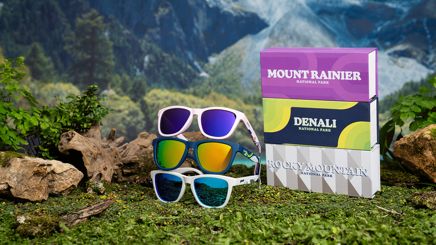 Three different special edition goodr National Park sunglasses stacked on top of each other (Mountain Rainier, Denali, and Rocky Mountain), on a mossy surface with mountains and trees in the background.