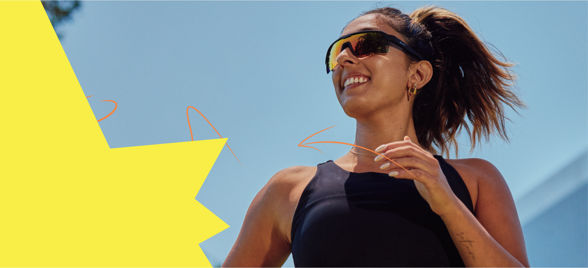 A woman is jogging outdoors under a clear blue sky, wearing reflective wraparound sunglasses and a black athletic top. Her hair is tied back in a high ponytail, and she has a joyful expression while running.