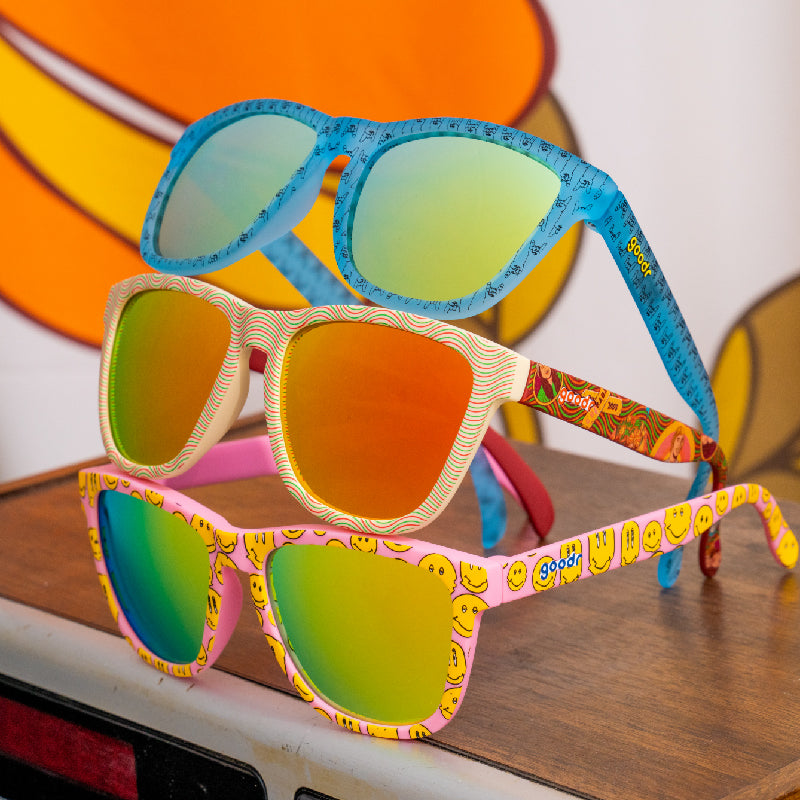 Three colorful, patterned sunglasses stacked on a table, with colorful artwork blurred in the background.
