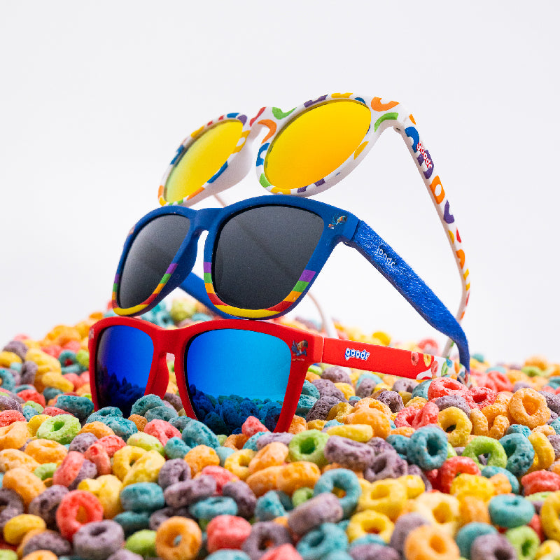 Three pairs of colorful sunglasses are stacked on a pile of Froot Loops cereal. The top pair has round white frames with yellow reflective lenses, the middle pair has blue frames with black lenses, and the bottom pair is red with blue mirrored lenses.