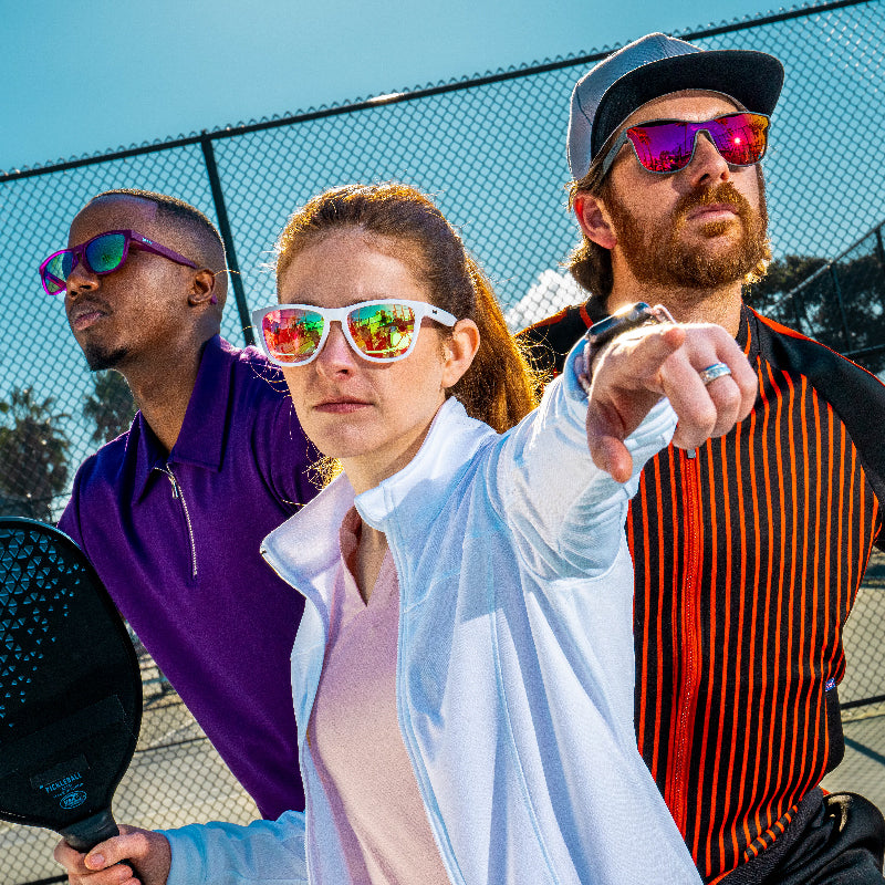 Three pickleball players in athletic clothing, wearing various, colorful styles of goodr sunglasses with reflective lenses.
