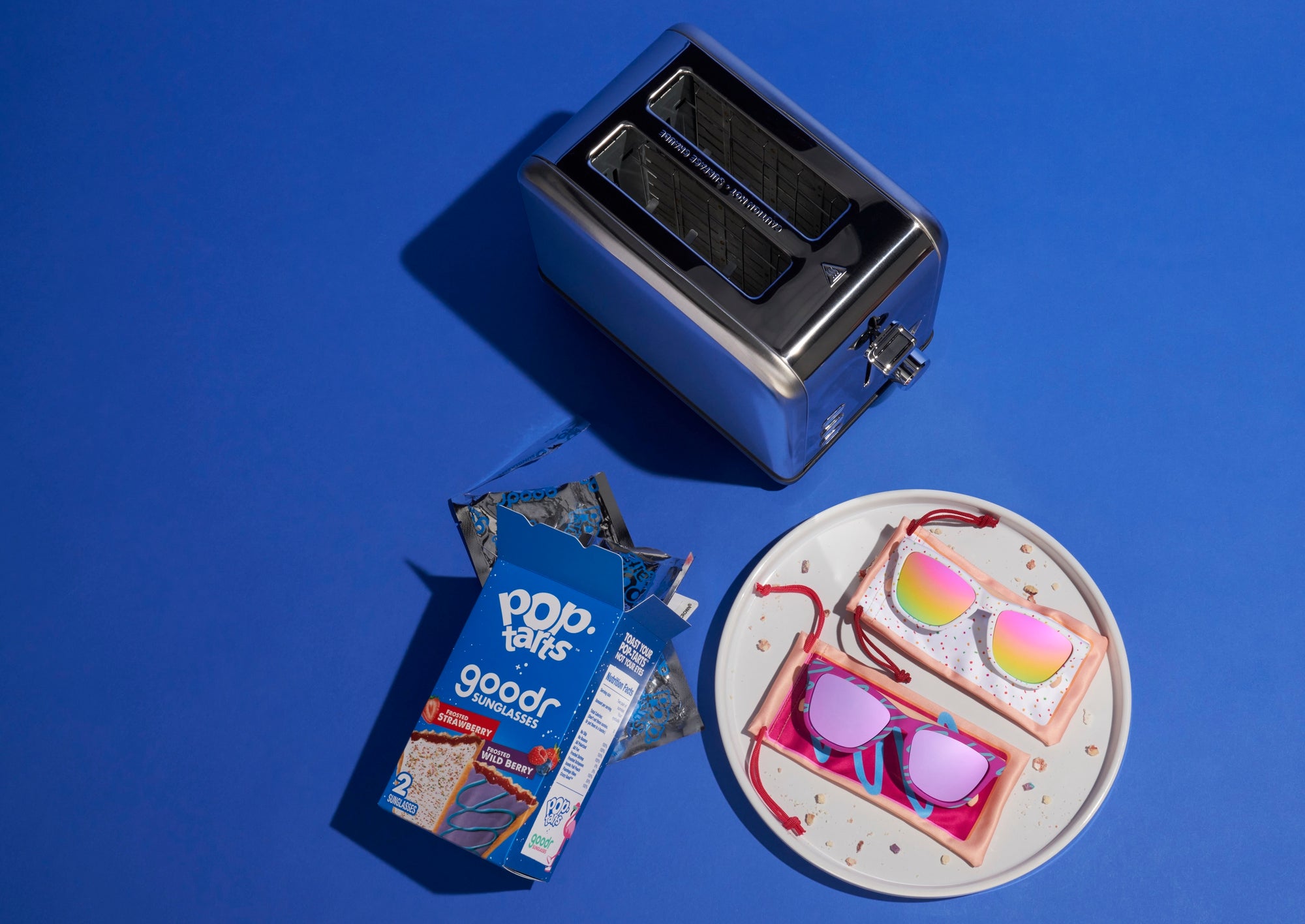 A playful flat-lay image featuring a collaboration between goodr sunglasses and Pop-Tarts. The scene shows a toaster, an open Pop-Tarts box, and a plate with two frosted Pop-Tarts on top. The Pop-Tarts are designed to look like sunglasses pouches, with colorful reflective Goodr sunglasses placed on top of them.