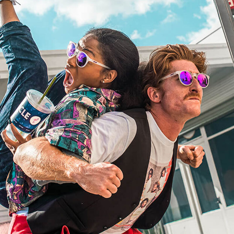 A playful scene of two people wearing mirrored purple "goodr" sunglasses. The man, with a mustache and vest, is carrying the woman on his back as she holds a large drink cup and reacts dramatically